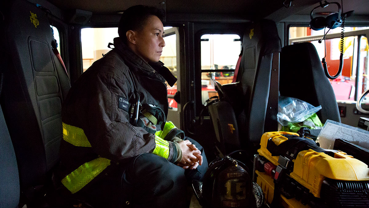 A woman firefighter sits in the cab of a firetruck in her jacket and gear.