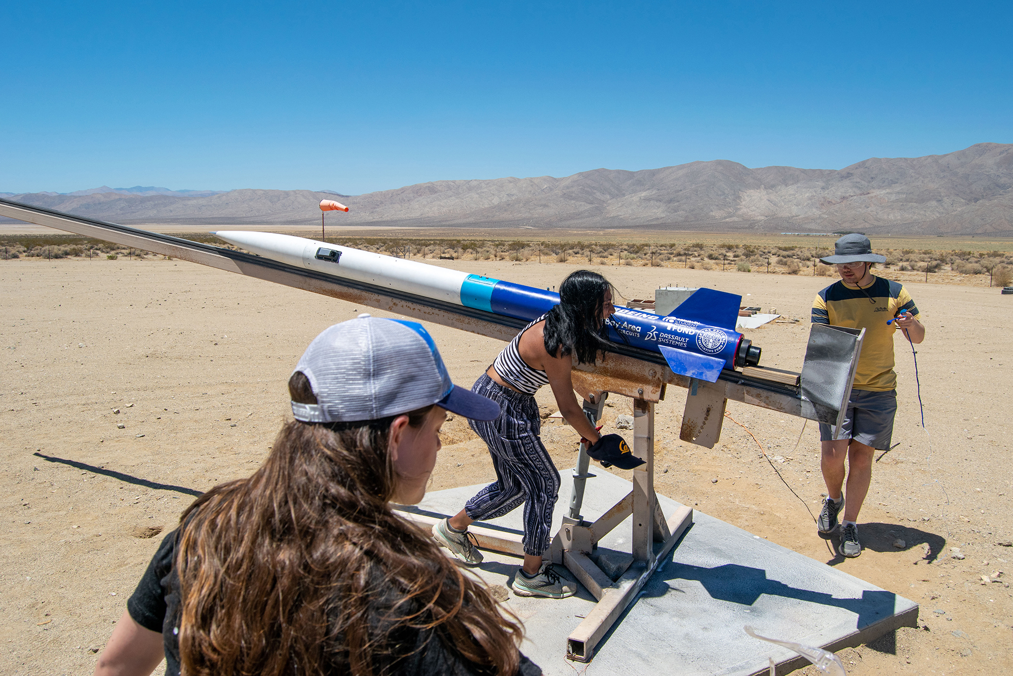 2 women and a man ready a 6-foot blue and white rocket for launch in the desert
