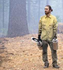 A photo of a man in protective fire gear holding two drip torches in the forests
