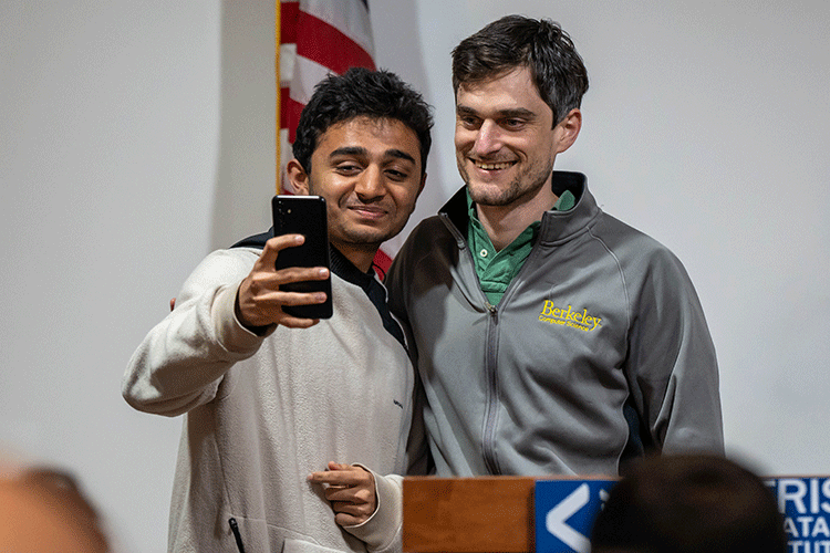 John Schulman stands beside a young person who is taking a selfie with their phone. 
