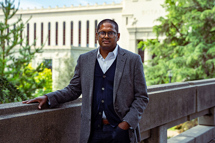 political science Professor Desmond Jagmohan standing on campus, wearing a tweed jacket over a cardigan and a light shirt