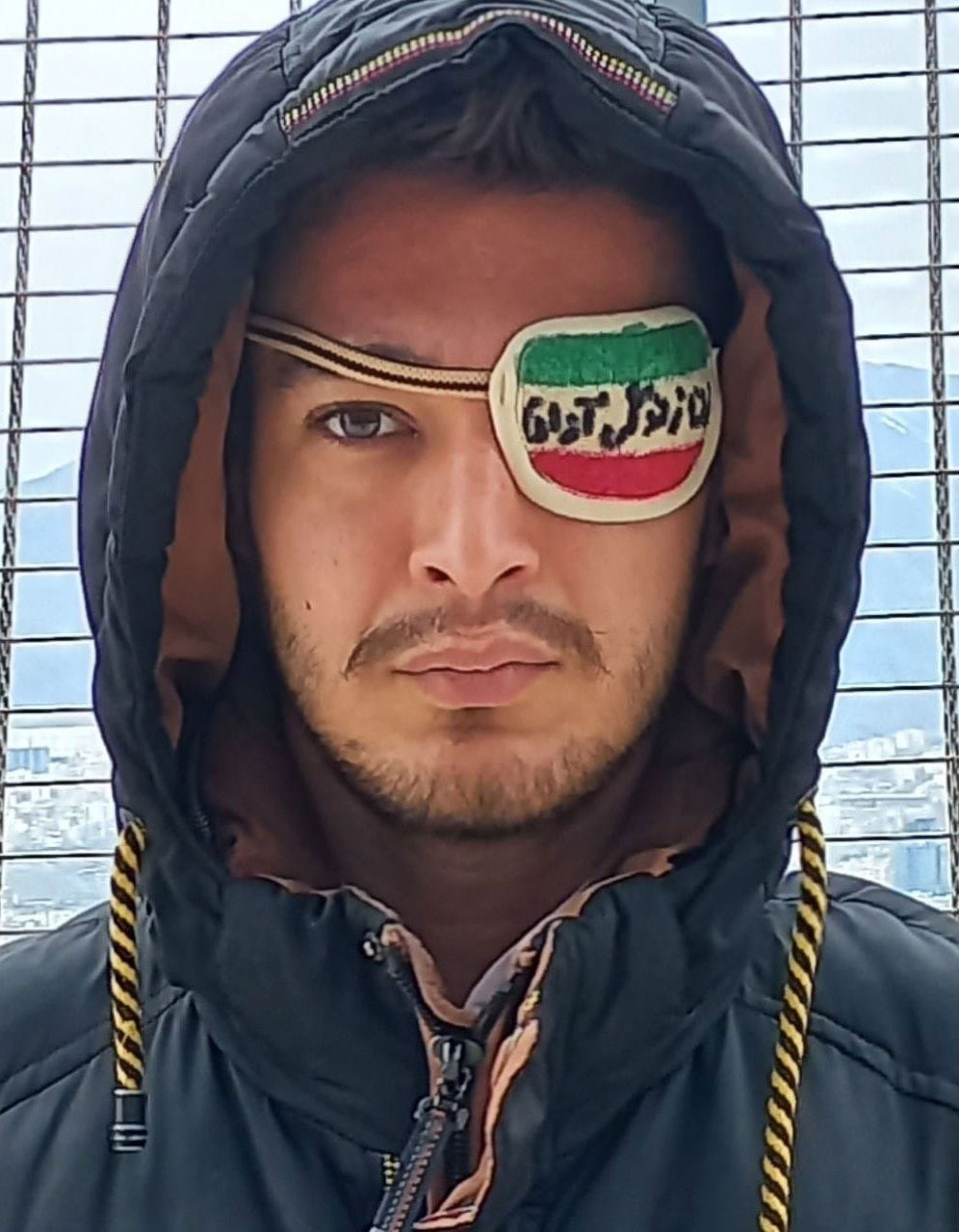 A portrait of Iranian dissident Hossein Noorinikoo with a patch over his blinded left eye. The patch bears the red, white and green bands of the Iranian flag, with script in Farsi. 