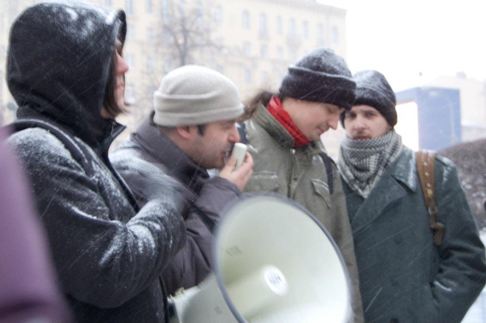 Four people stand in the snow while one of them with a light-colored beanie speaks into a microphone