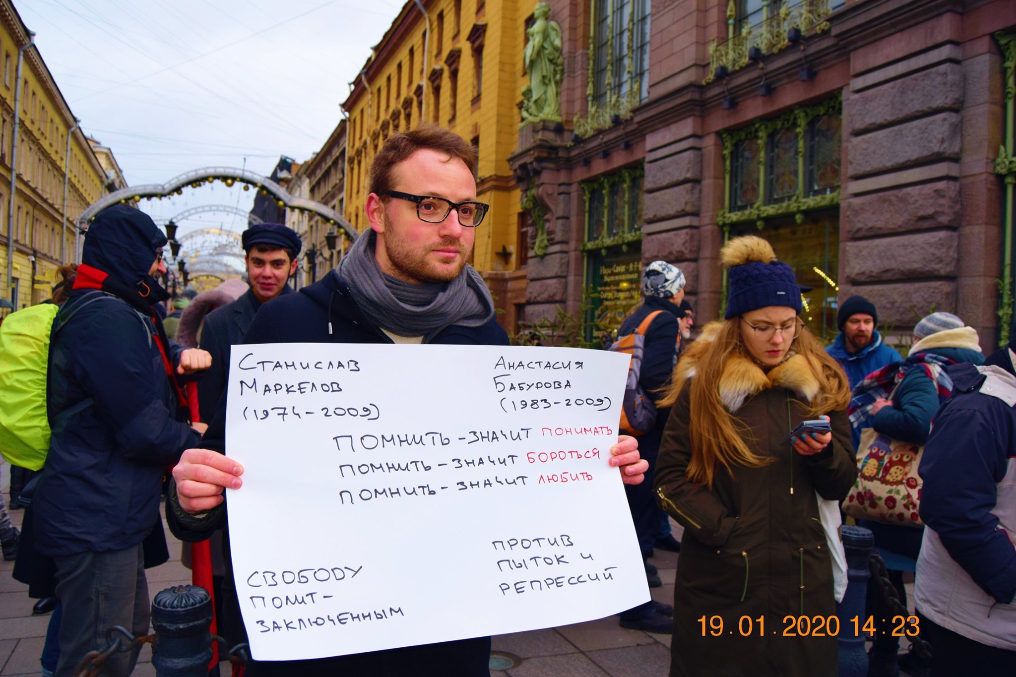 A person with glasses and a scarf hold a white cardboard sign with Russian writing