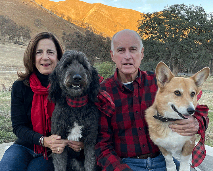 A photo shows Jerry and Anne Brown sitting outside with a black dog and a tan dog. Grassy hills are in the background.