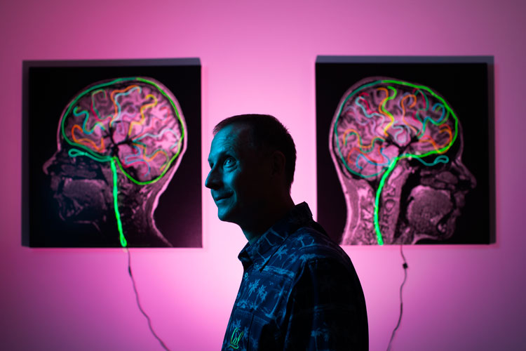 a picture of a person standing to the side and looking toward the left in a dimly lit room in front of two images of brain scans on a pink lit wall