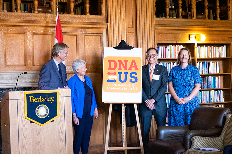Robbert Dijkgraaf, the Dutch minister of education, culture and science; Chancellor Christ; Stanford professor Guido Imbens; and Renske Heemskerk, science and education attaché at the Netherlands Embassy in Washington, D.C., pose for photos in Morrison Library after the announcement of DNA-US. There is a large sign saying DNA-US on an easel. 