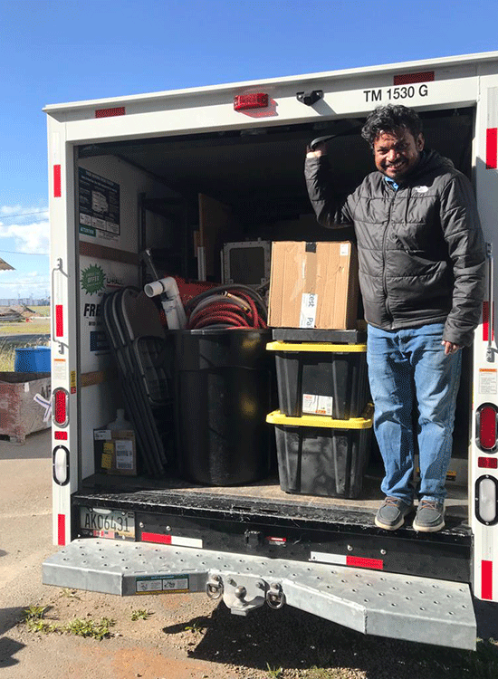 On a bright sunny day, a person stands next to a stack of boxed equipment inside the open end of a U-Haul truck.