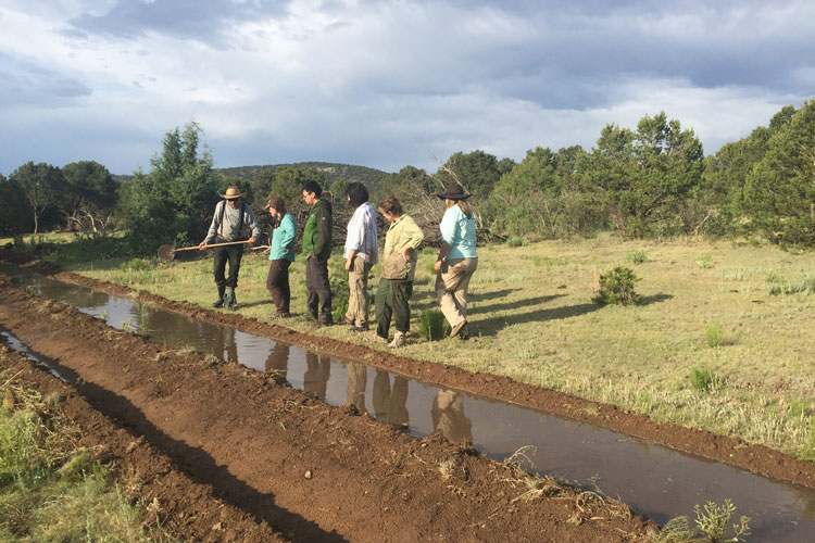 a team of people stand next to a irrigation ditch under a cloudy sky