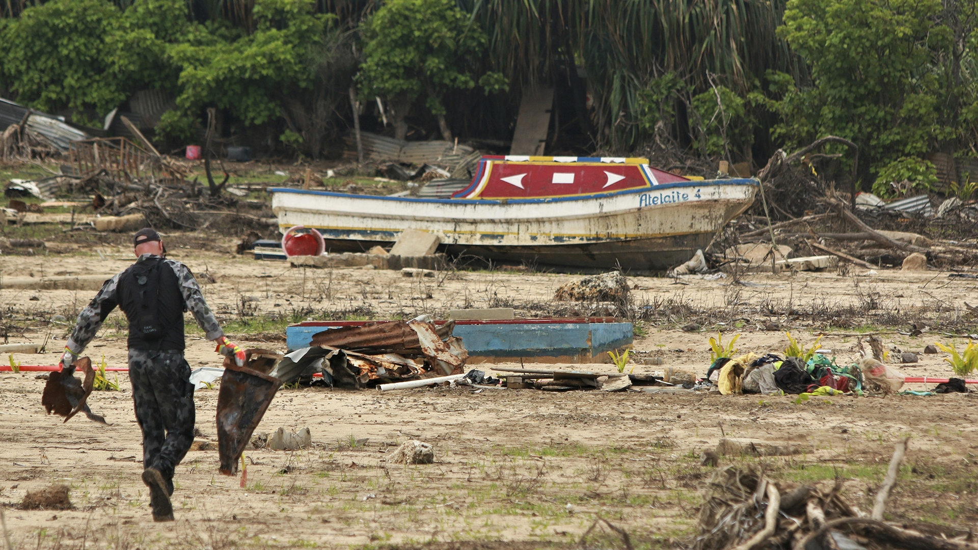 a person cleans up debris on the island of Tonga after a volcanic eruption in 2022 caused a tsunami. A boat is on land.
