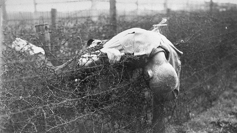 A grainy black and white photo features a corpse caught in barbed wire at Leipzig-Thekla, a sub-camp of the Nazi Buchenwald death camp. 