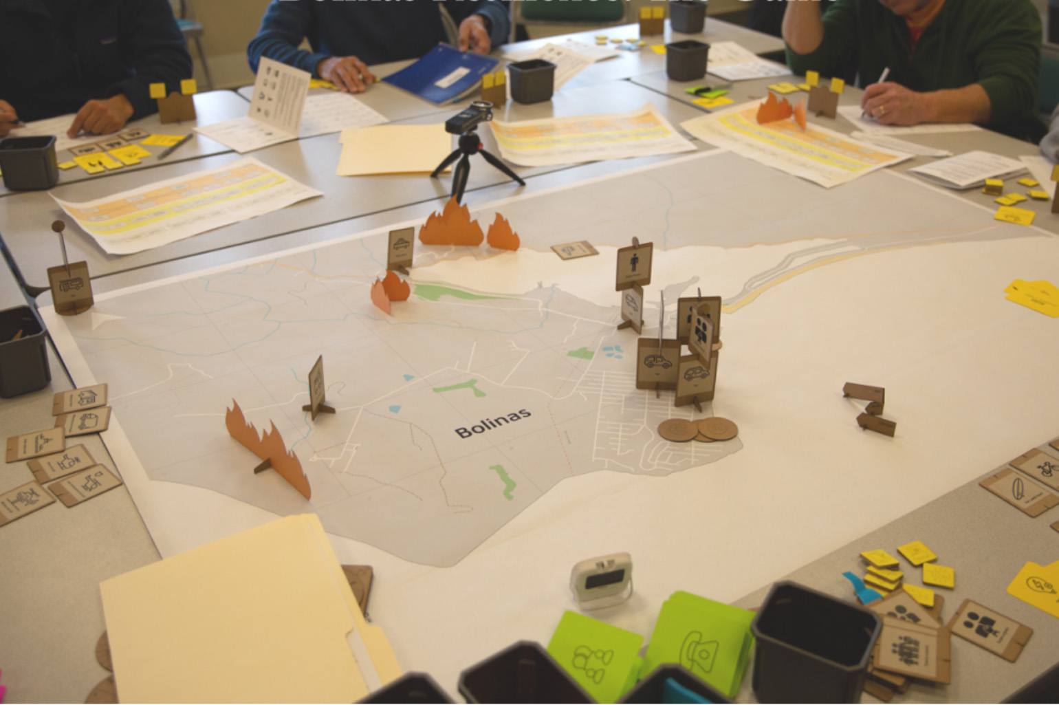 A board game on a conference room table. The game space, a map of Bolinas, California, is printed on a large piece of paper. Cardboard game pieces are placed around the map, with squares labeled to indicate individual vehicles and flame shapes indicating wildfire. Scattered on the table around the board are notebooks, pens and game cards. At the top and right edges are three people with only their hands visible. They appear to be writing notes, flipping through notebooks and looking at game pieces.