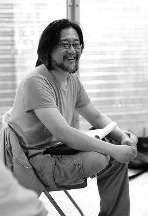 Stan Lai sitting in a chair with writing pad in lap, smiling.