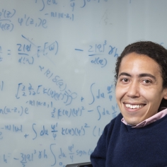 Professor Ahmad Omar sitting in front of a glass whiteboard marked with equations related to his research. 
