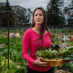 Elizabeth standing in the Gill Tract Community Garden wearing a red shirt holding a basket of produce