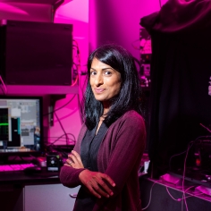 Teresa Puthussery in lab setting with pink filtered lighting and machinery in background.