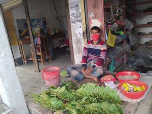 A photo of a Bangladeshi shop keeper wearing a red cloth face mask, while sitting outside behind a selection of produce.