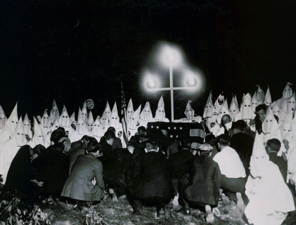 In a haunted nighttime a century ago, hooded members of the Ku Klux Klan kneel with new recruits before an American flag and a torch-lit cross.