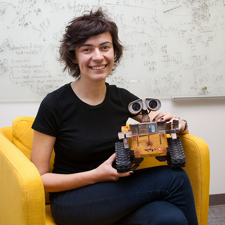 Associate Professor Anca Dragan holds a small yellow robot while sitting in a bright yellow chair.