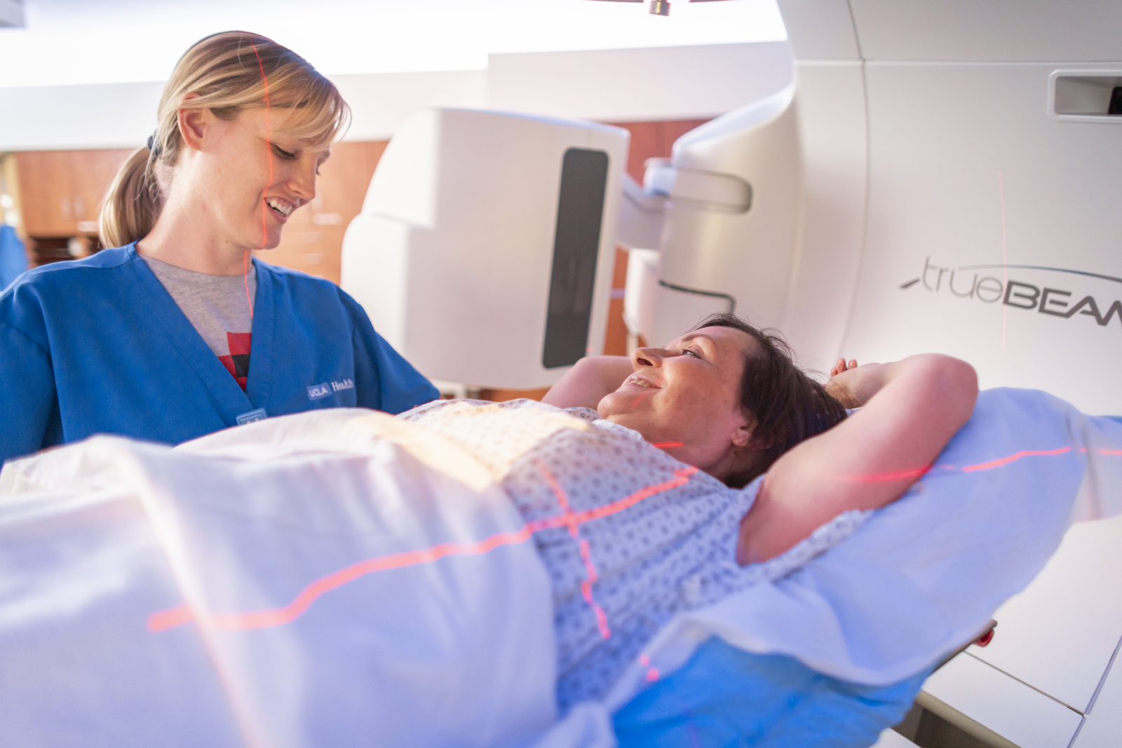 A woman in a medical gown lies on her back inside a diagnostic machine, smiling at a technician preparing to take an image.