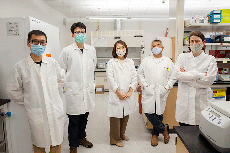 A photo shows five individuals standing in a biology lab. Each person is wearing a lab coats and a face mask.