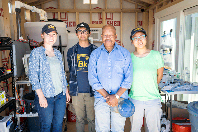 Four people pose for a photo inside a shed that is filled with scientific equipment.