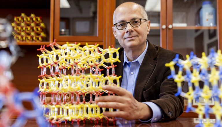 a seated man in jacket and blue shirt holding a large yellow and red molecular structure