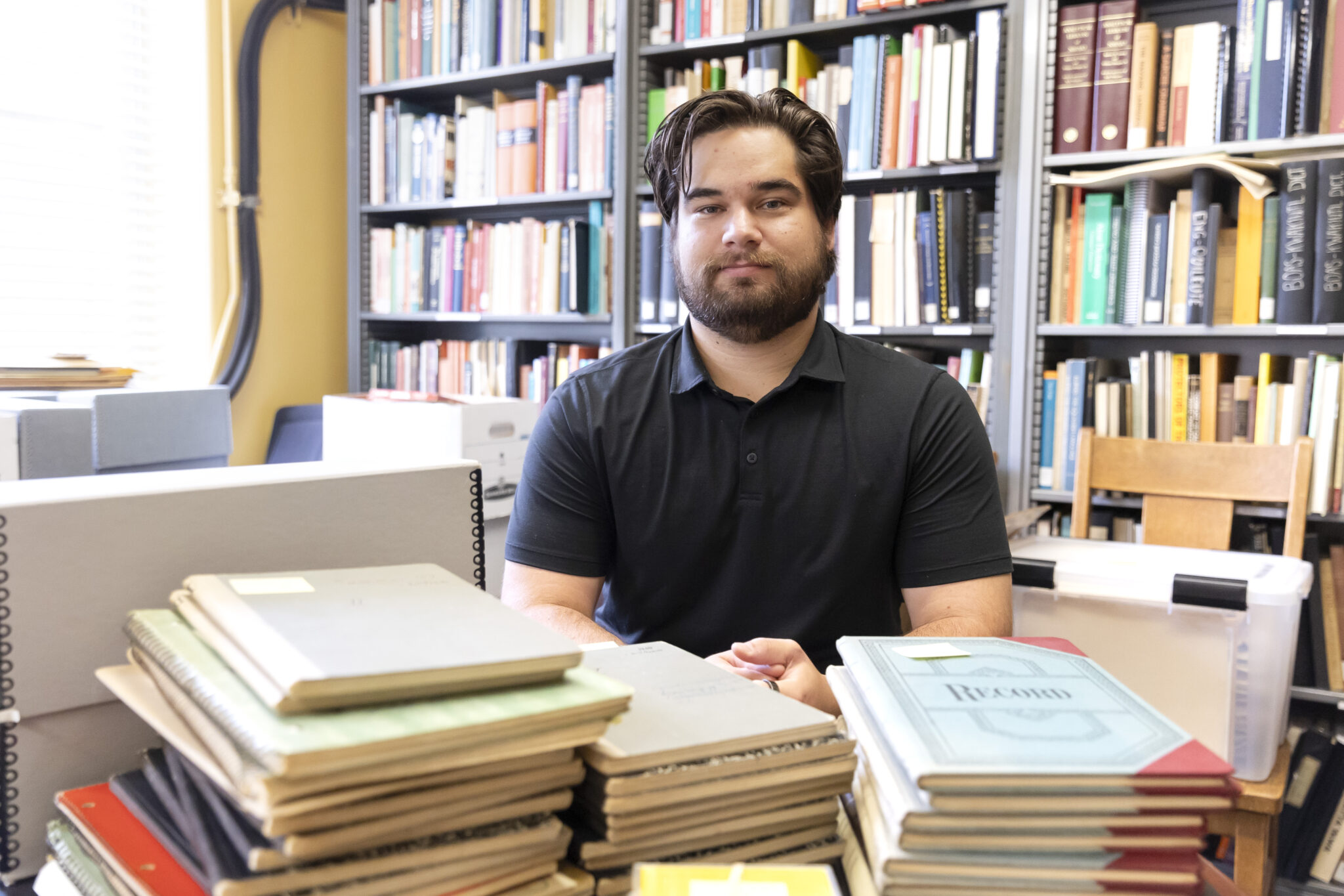 Jonathan Cirelli seated at a table, with dozens of notebooks from the California Language Archive stacked in front of him and a bookshelf in the background.