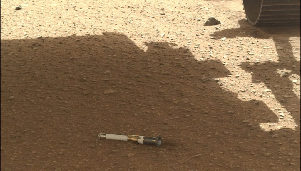 a small white and black tube sits on pebbly red ground in the shade of the Insight lander on Mars