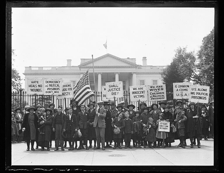 In a black and white photograph, a group of people, including children, stand in front of the White House holding signs that say "shall justice die?" and others