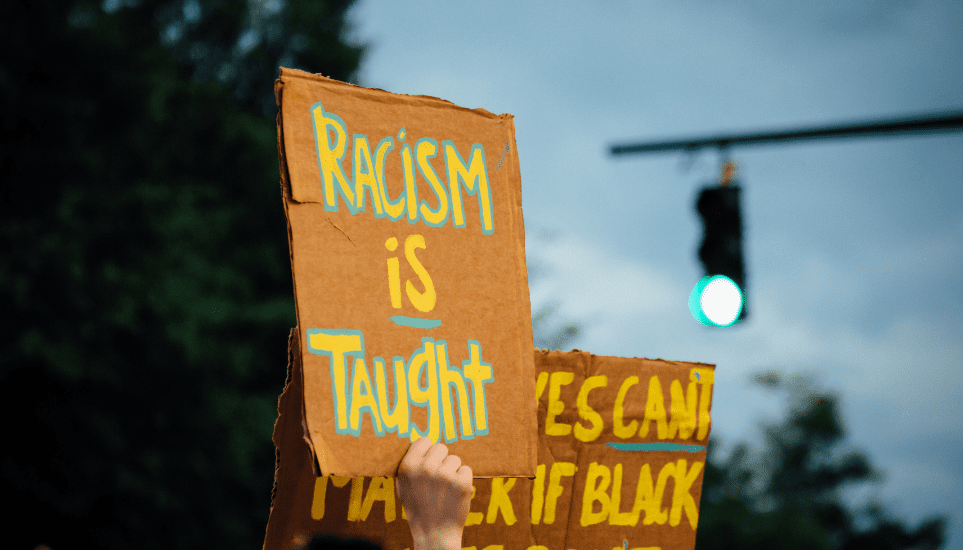 Hand holding a sign that reads "Racism is Taught"