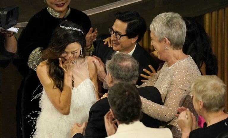 Michelle Yeoh stands with a hand to her mouth as Jamie Lee Curtis and Ke Huy Quan congratulate her.