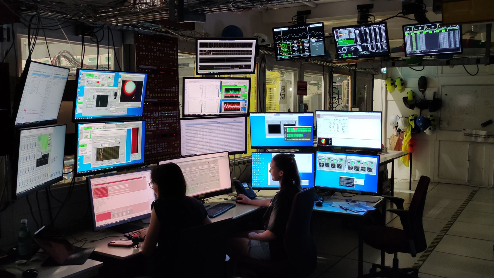 two women sitting in dark control room in front of 16 computer monitors