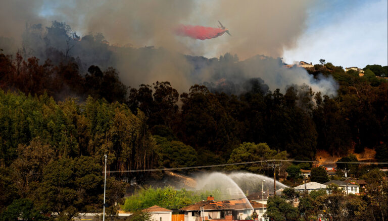 Smoke emerges from forested hills. In the foreground, two fire trucks aim streams of water at a vulnerable home. Overhead, an aircraft releases a plume of red flame retardant chemicals.