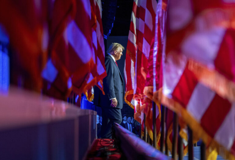 With a row of American flags on the left and right side of the frame, Donald Trump walks in the distance while supporters cheer