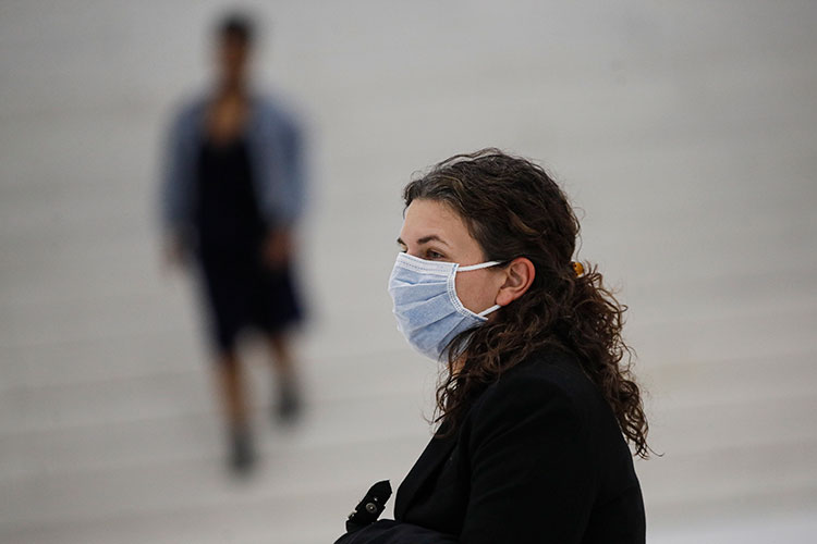 A woman stands on a street wearing a light blue surgical mask.