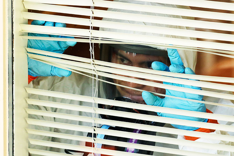 A man wearing a protective suit, gas mask, and blue gloves peers through a set of window blinds