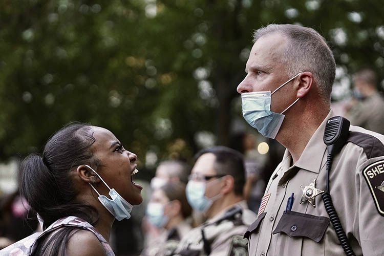 a black woman with a mask at her chin yells at a white male police officer wearing a mask and staring into the distance