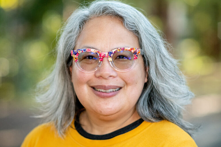 Abigail De Kosnik, an associate professor of theater, dance, and performance studies and a 2023 Distinguished Teaching Award winner, smiles at the camera. She had shoulder-length gray hair and colorful eyeglasses.