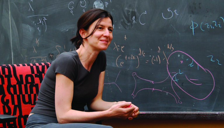 a woman in a black T-shirt sitting in front of a blackboard talking to someone off-camera