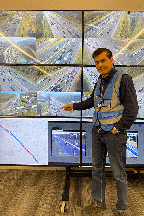 A researcher wearing a blue safety vest points to an image of traffic on a large monitor.