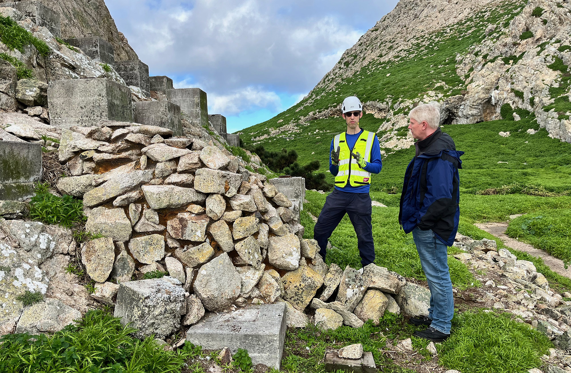 man in white hat and yellow vest talks with Richard Allen in front of pile of rocks covering seismic sensors