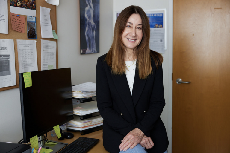 Annamaria Bellezza, a distinguished senior lecturer in Italian, sits on the edge of a desk with her hands folded across her lap. She has long brown hair and is wearing a dark blazed and jeans.