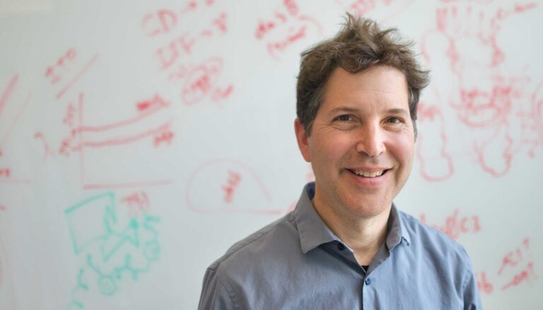 a man in blue shirt with tousled hair stands in front of a whiteboard