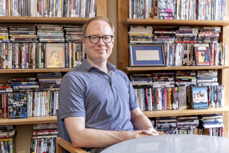 A person wearing a polo shirt and glasses sits at a table in front of a large bookshelf that is filled with DVDS.