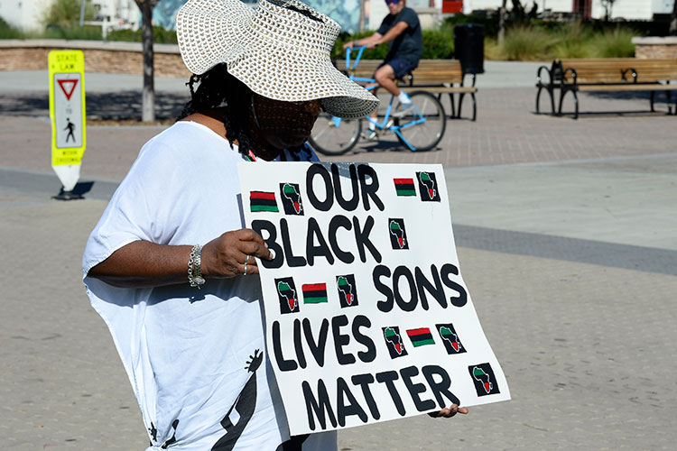 A Black woman stands in the street, wearing a large hat that obscures her face. She holds a sign that says "Our Black sons lives matter."