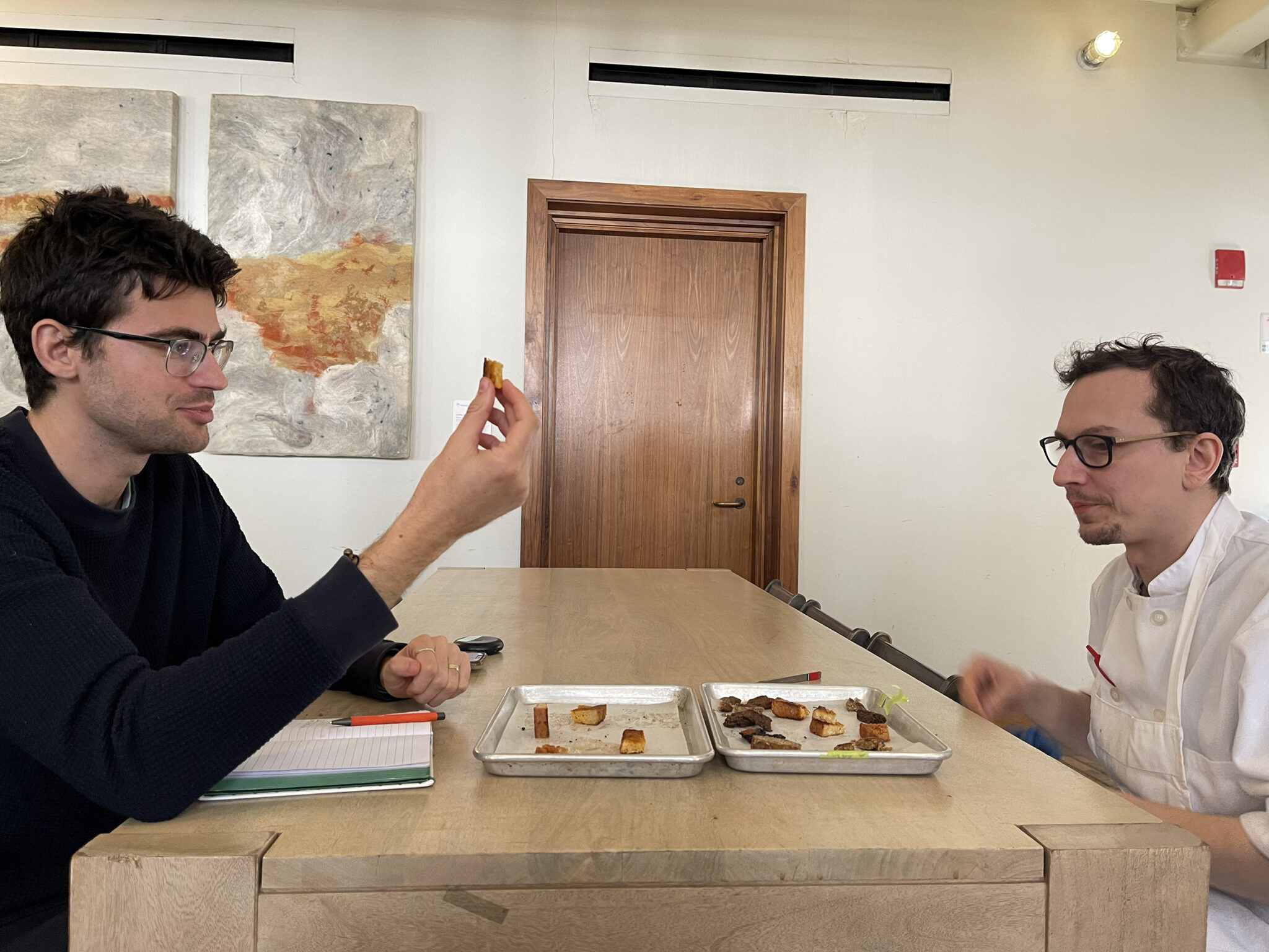 two men, one in white chef's clothing, tasting small bits of food sitting on metal trays