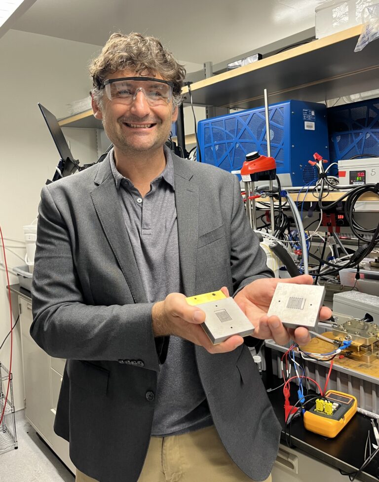 man in goggles standing in a lab holding 2 devices in his hands