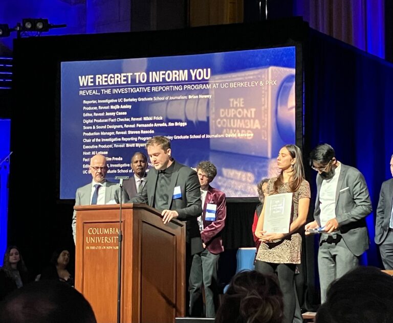 A group of people on an awards stage watch a man giving an acceptance speech.