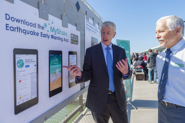 Richard Allen, in a suit, points to a large printout of smartphone screens showing how the MyShake app works.